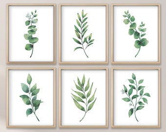 May include: Six framed botanical prints featuring watercolor illustrations of green leaves and branches. The prints are displayed on a light-colored wall, above a woven chair and a bed with a beige blanket.