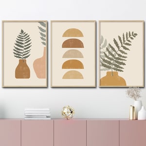 May include: Set of three minimalist art prints featuring abstract shapes and fern leaves in shades of brown, green, and beige. The prints are framed in a light wood frame and are displayed on a pink wall.