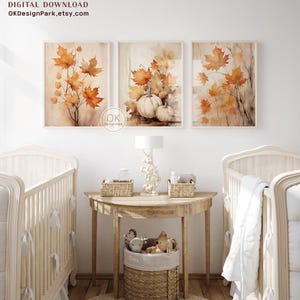 May include: Three watercolor prints featuring fall leaves and pumpkins. The prints are framed in a light wood frame and hung on a white wall. The prints are displayed in a nursery setting with two cribs and a side table with a lamp and a basket of toys.