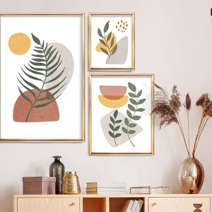 Puede incluir: Tres impresiones de arte abstracto con un dise&ntilde;o minimalista que presenta hojas verdes, c&iacute;rculos amarillos y naranjas y formas marrones y beige. Las impresiones est&aacute;n enmarcadas en oro y colgadas en una pared blanca.