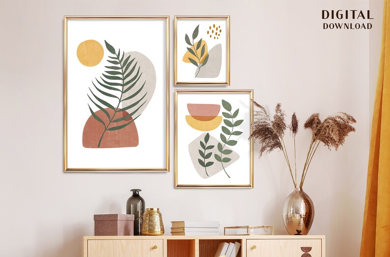 Puede incluir: Tres impresiones de arte abstracto con un dise&ntilde;o minimalista que presenta hojas verdes, c&iacute;rculos amarillos y naranjas y formas marrones y beige. Las impresiones est&aacute;n enmarcadas en oro y colgadas en una pared blanca.