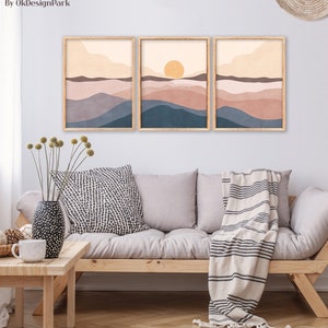 May include: Three framed landscape art prints with a minimalist design. The artwork features layered hills in shades of blue, brown, and beige, with a yellow sun in the center print. The frames are light brown. The text "Instant Download Art - On Etsy By OkDesignPark" is at the top.