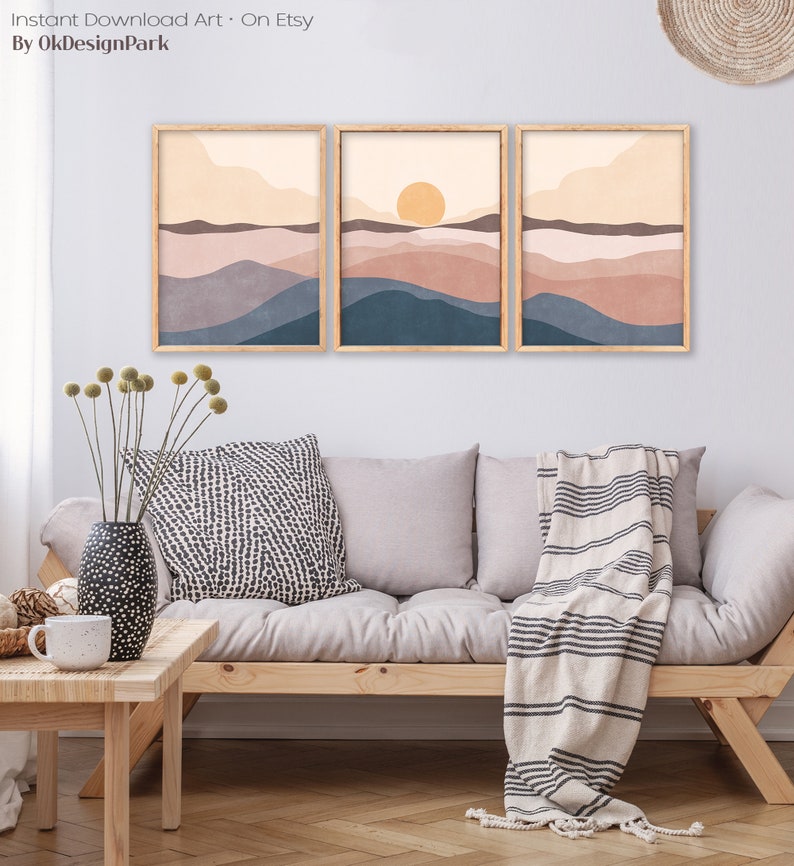 May include: Three framed landscape art prints with a minimalist design. The artwork features layered hills in shades of blue, brown, and beige, with a yellow sun in the center print. The frames are light brown. The text "Instant Download Art - On Etsy By OkDesignPark" is at the top.