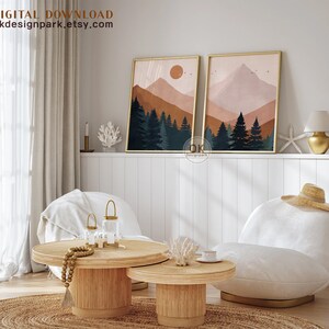 May include: Two abstract landscape prints with a mountain range, trees, and a sun in the sky. The prints are in a gold frame and are hanging on a white wall. The prints are above a white couch with a straw hat on it.