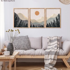 K&ouml;nnte beinhalten: Drei gerahmte Drucke einer minimalistischen Landschaft mit Bergen, B&auml;umen und einer Sonne. Die Drucke sind in einem hellbraunen Rahmen und h&auml;ngen an einer wei&szlig;en Wand &uuml;ber einem hellgrauen Sofa.