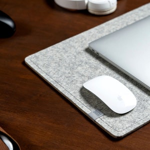 Peut inclure: Un tapis de bureau en feutre gris avec un ordinateur portable, une souris sans fil blanche et un étui en cuir marron sur une surface en bois.