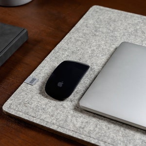 Könnte beinhalten: Eine graue Filzunterlage für den Schreibtisch mit einer schwarzen kabellosen Maus und einem silbernen Laptop. Der Laptop hat ein Apple-Logo auf der Rückseite.