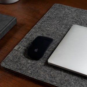Puede incluir: Una alfombrilla de escritorio de fieltro gris con un ordenador portátil, un ratón inalámbrico negro y un cuaderno negro sobre una superficie de madera.