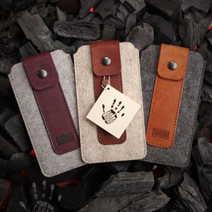 May include: Three felt phone cases with leather accents. The cases are gray, beige, and brown. Each case has a leather flap with the word "CHARISMA" embossed on it. The beige case has a small white tag with a handprint on it.