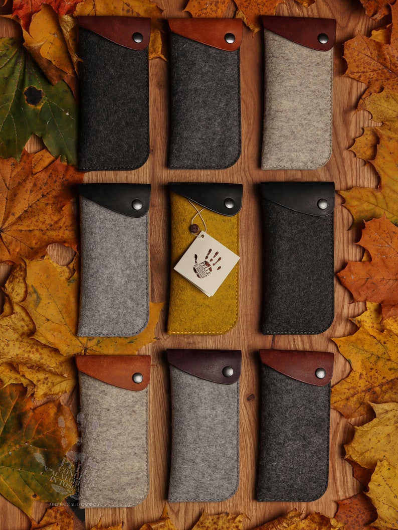 May include: Ten felt eyeglass cases in various colors, including gray, brown, and beige. The cases have a leather flap with a button closure. The cases are arranged in three rows on a wooden surface with autumn leaves scattered around them.