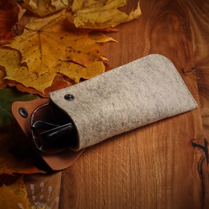 May include: A gray felt eyeglasses case with a brown leather flap and a snap closure. The case is lying on a wooden surface with autumn leaves scattered around it.