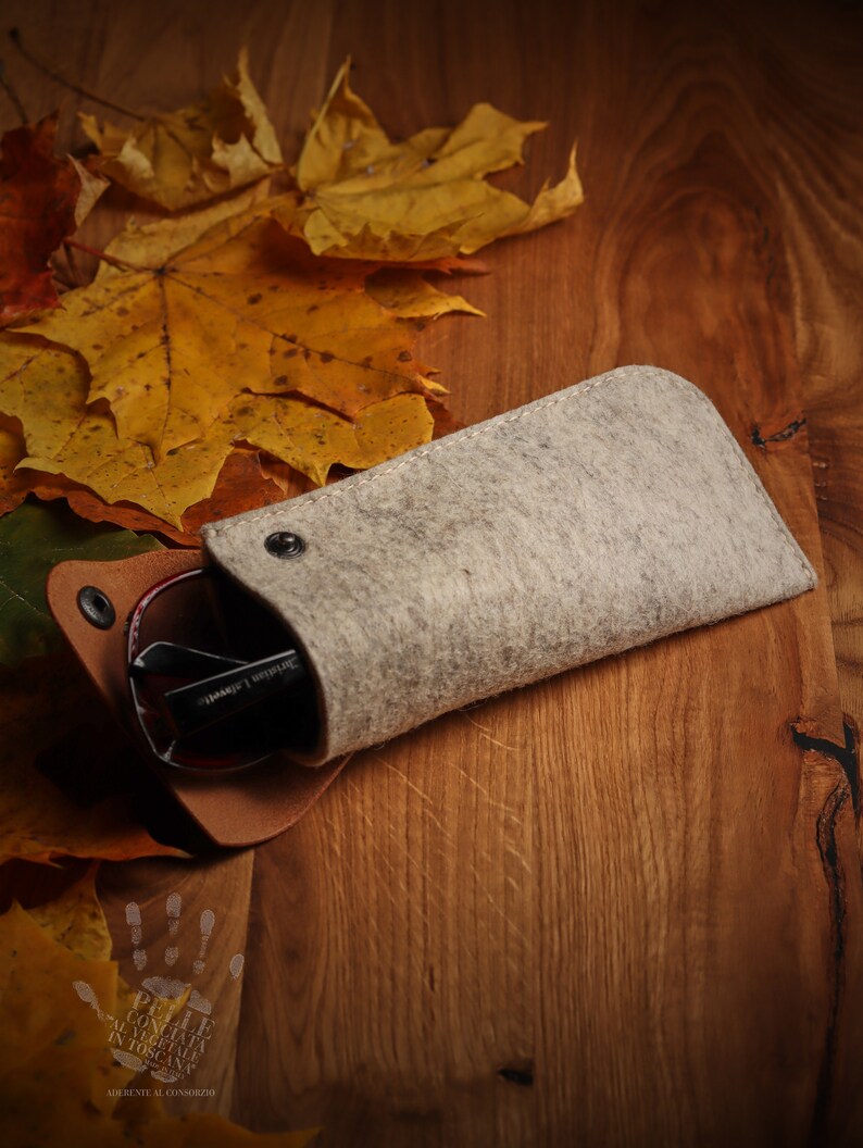 May include: A gray felt eyeglasses case with a brown leather flap and a snap closure. The case is lying on a wooden surface with autumn leaves scattered around it.