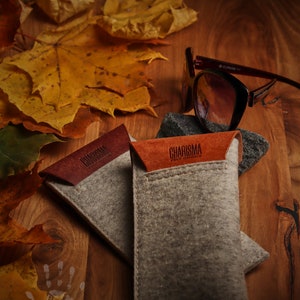 May include: Two gray felt eyeglass cases with brown leather accents. The cases have the text "CHARISMA PELLETTERIA" embossed on the leather. The cases are on a wooden surface with autumn leaves.