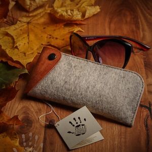 May include: A gray felt glasses case with a brown leather flap and a snap closure. The case is sitting on a wooden surface with autumn leaves and a pair of sunglasses. The case has a tag attached with the text "PELLE CONCIATA AL VEGETALE IN TOSCANA" and a handprint logo.  ADERENTE AL CONSORZIO.