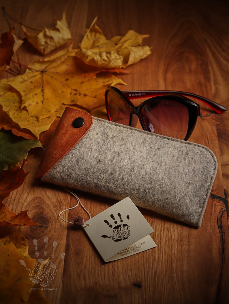 May include: A gray felt glasses case with a brown leather flap and a snap closure. The case is sitting on a wooden surface with autumn leaves and a pair of sunglasses. The case has a tag attached with the text "PELLE CONCIATA AL VEGETALE IN TOSCANA" and a handprint logo.  ADERENTE AL CONSORZIO.