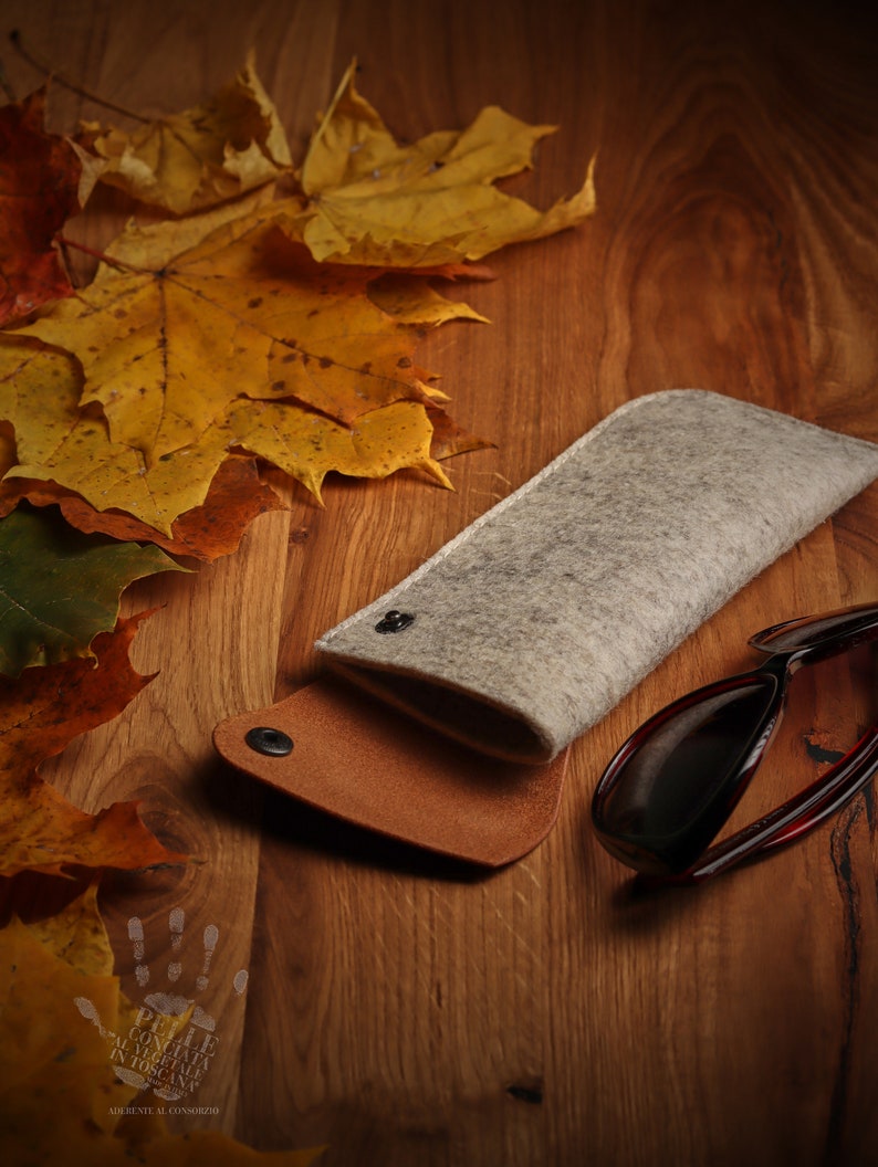 May include: A brown leather and gray felt glasses case with a snap closure. The case is lying on a wooden surface with autumn leaves scattered around it. A pair of dark sunglasses is also on the surface.