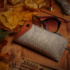 May include: A gray felt eyeglasses case with a brown leather flap and a snap closure. The case is lying on a wooden surface with autumn leaves and a wooden stamp.