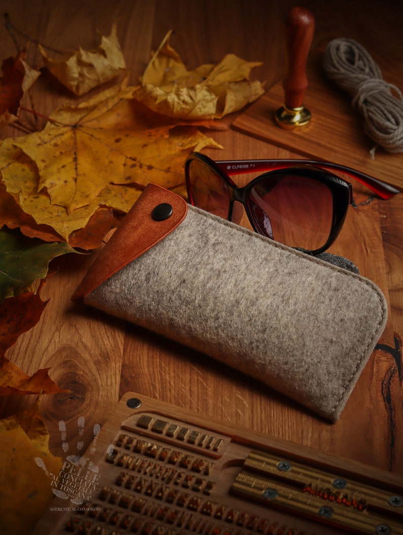 May include: A gray felt eyeglasses case with a brown leather flap and a snap closure. The case is lying on a wooden surface with autumn leaves and a wooden stamp.