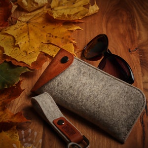 May include: A gray felt eyeglasses case with a brown leather flap and a brown leather keychain with the word "remarkable" on it. The case is sitting on a wooden surface with autumn leaves.