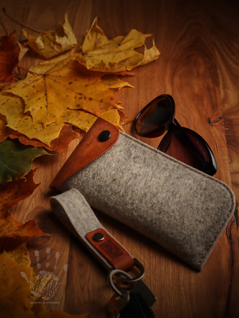 May include: A gray felt eyeglasses case with a brown leather flap and a brown leather keychain with the word "remarkable" on it. The case is sitting on a wooden surface with autumn leaves.