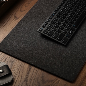Puede incluir: Un teclado inalámbrico negro con una alfombrilla de escritorio de fieltro gris sobre una superficie de madera.