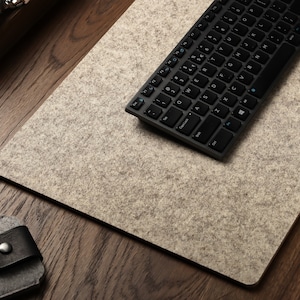 May include: A gray felt desk mat with a black wireless keyboard on top. The keyboard has a standard layout with black keys and white lettering.