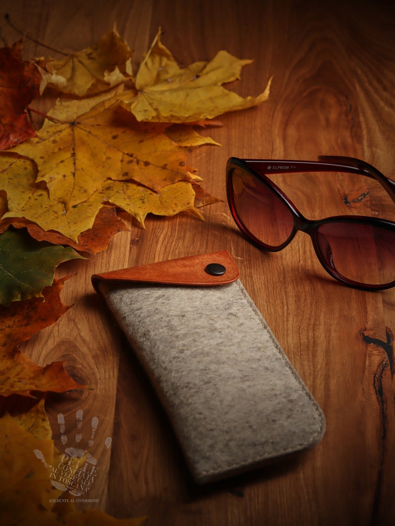 May include: A gray felt eyeglasses case with a brown leather flap and a snap closure. The case is lying on a wooden surface with autumn leaves and a pair of brown sunglasses.