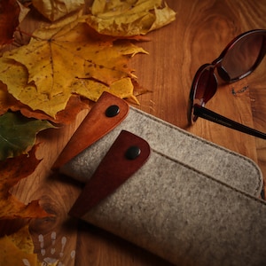 May include: A gray felt glasses case with brown leather accents and a snap closure. The case is lying on a wooden surface with autumn leaves scattered around it. A pair of brown sunglasses with the text "Stefano Latterini" on the frame is lying next to the case.