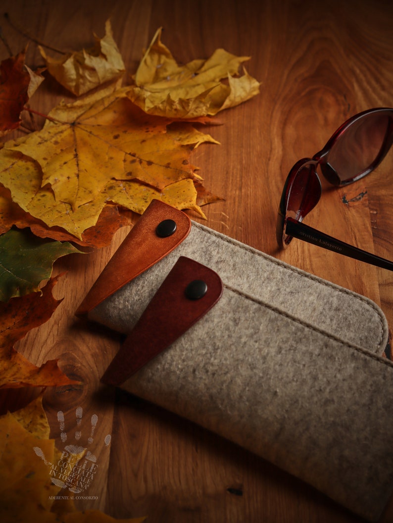 May include: A gray felt glasses case with brown leather accents and a snap closure. The case is lying on a wooden surface with autumn leaves scattered around it. A pair of brown sunglasses with the text "Stefano Latterini" on the frame is lying next to the case.