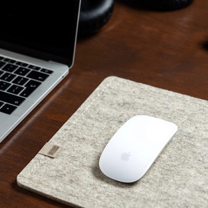 Puede incluir: Un ratón inalámbrico blanco con un logotipo de Apple en una alfombrilla de ratón de fieltro gris. La alfombrilla de ratón está sobre un escritorio de madera.
