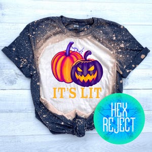 May include: A black and white t-shirt with a Halloween theme. The shirt features a graphic of two pumpkins, one orange and one purple, with the text "IT'S LIT" in orange. The shirt has a bleached design.