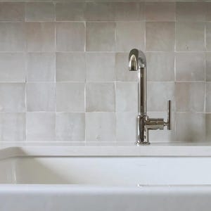 Op de afbeelding: Een witte keukenwasbak in boerderijstijl met een chromen kraan. De wasbak is rechthoekig met afgeronde randen en staat onder een wit aanrechtblad. De achtergrond heeft vierkante, lichtgekleurde tegels. De kasten eronder zijn grijs met zilveren knoppen.