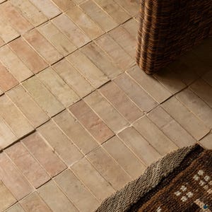 May include: A close-up shot of a floor tiled with rectangular, light brown bricks arranged diagonally. A woven wicker chair leg and a patterned rug are visible, adding texture and depth to the scene.