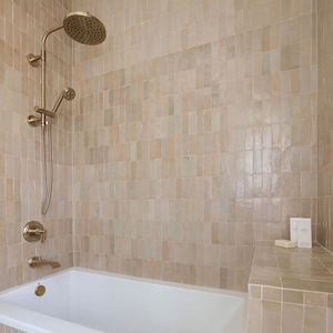 Puede incluir: Un baño moderno con una bañera blanca y paredes de azulejos beige. Un cabezal de ducha y un grifo de color dorado están montados en la pared. El cabezal de la ducha es redondo y el grifo tiene un mango de palanca. El baño tiene un banco incorporado.