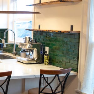 Könnte beinhalten: Eine Küche mit einer Espressomaschine aus Edelstahl auf einer weißen Arbeitsplatte. Der Fliesenspiegel besteht aus grünen Ziegeln. Holzregale sind darüber angebracht. Zwei Stühle mit dunklen Metallrahmen und braunen Sitzen befinden sich im Vordergrund.
