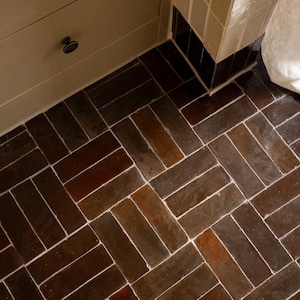 Può includere: Pavimento a spina di pesce con piastrelle rettangolari marrone scuro. Le piastrelle sono disposte in diagonale, con linee di fuga bianche che le separano. Il pavimento è accanto a un mobile beige e a una parete piastrellata.