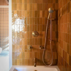 Könnte beinhalten: Eine Badezimmerdusche mit einem goldfarbenen Duschkopf, Wasserhahn und Armaturen. Die Duschwand ist mit rechteckigen, braunen Fliesen bedeckt. Die Badewanne ist weiß.
