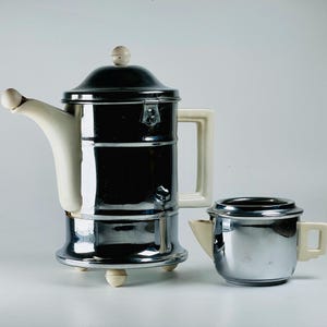 Könnte beinhalten: Ein chromfarbenes und weißes Keramik-Teekannen- und Milchkännchen-Set. Die Teekanne hat einen runden Deckel mit einem weißen Knopf und einem weißen Ausguss. Das Milchkännchen hat einen weißen Griff und einen weißen Ausguss.