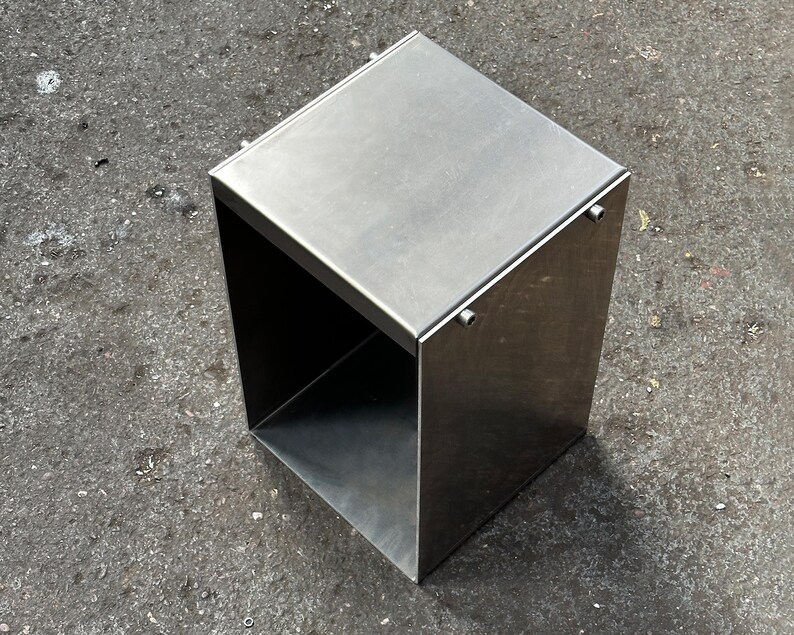 May include: A silver metal stool with a square top and open sides. The stool is made of metal and has a smooth, polished finish.