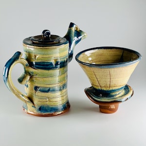 Op de afbeelding: Een handgemaakte keramische theepot en filterset met een blauw-groen gestreept ontwerp. De theepot heeft een unieke tuit en een deksel. Het filter is kegelvormig met een brede basis.