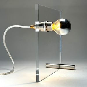 May include: A modern table lamp with a clear acrylic base and a silver metal shade. The lamp has a single light bulb with a silver finish.