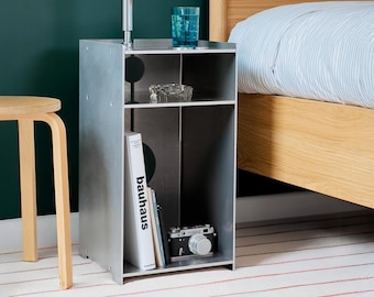 May include: A silver metal side table with three shelves. The top shelf has a glass ashtray and a blue glass. The middle shelf has a book titled "bauhaus" and a smaller book. The bottom shelf has a camera. The table is next to a bed with a white duvet cover.