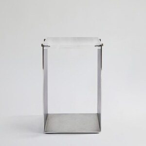 May include: A small, square, silver metal table with a clear acrylic top. The table has a simple, modern design.