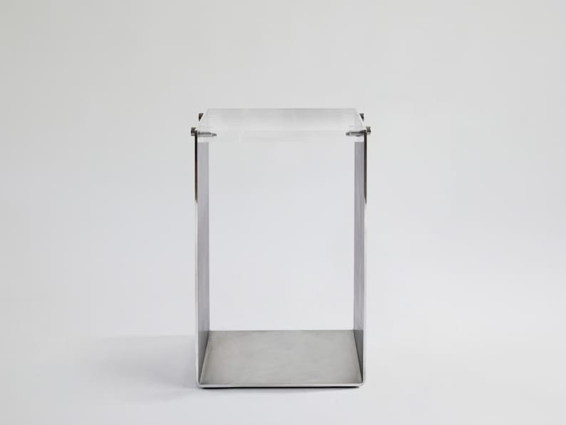 May include: A small, square, silver metal table with a clear acrylic top. The table has a simple, modern design.