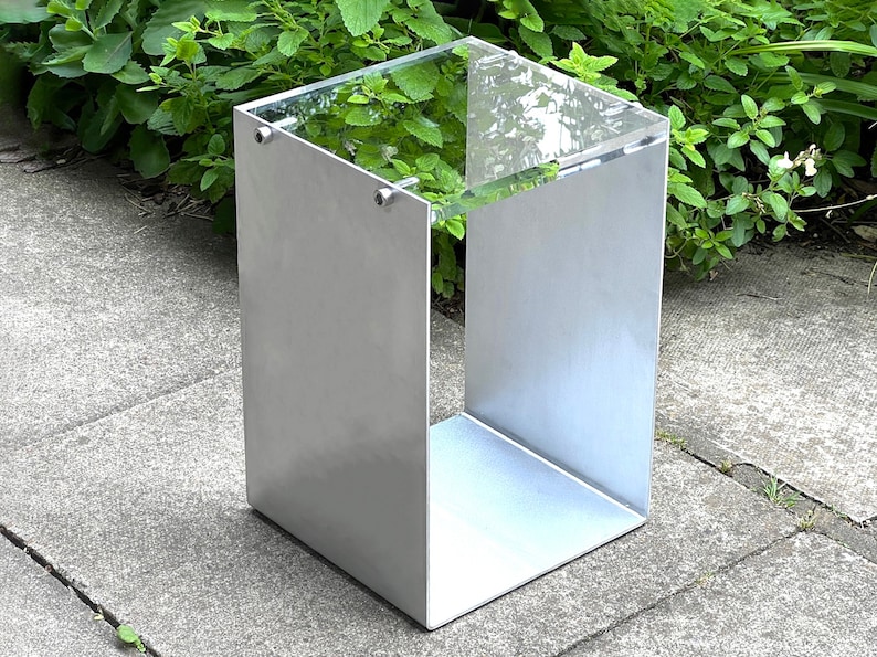 May include: A silver metal side table with a clear glass top. The table has a square base and a simple, modern design.