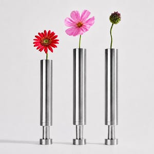 May include: Three cylindrical metal vases with a brushed finish, each featuring a narrow base and a wider top section. A red gerbera daisy, a pink cosmos flower, and a small green bud are placed individually in each of the three vertical vessels.