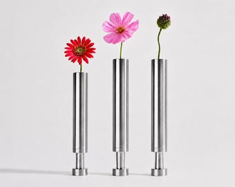 May include: Three cylindrical metal vases with a brushed finish, each featuring a narrow base and a wider top section. A red gerbera daisy, a pink cosmos flower, and a small green bud are placed individually in each of the three vertical vessels.