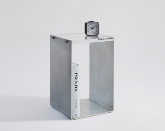 May include: A gray concrete side table with a glass top and a single shelf. A black alarm clock and a white book titled "PRADA CATWALK" are on the table.
