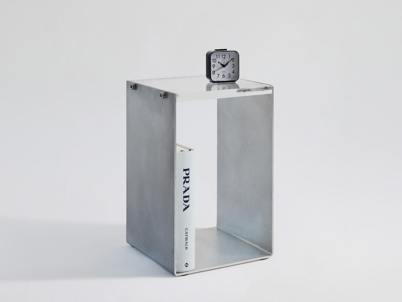 May include: A gray concrete side table with a glass top and a single shelf. A black alarm clock and a white book titled "PRADA CATWALK" are on the table.