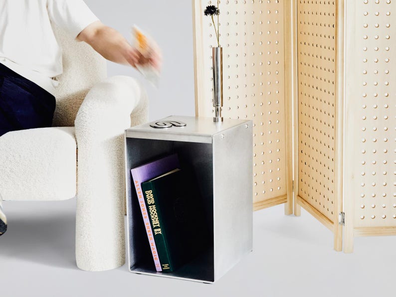 May include: A silver metal side table with a shelf and a book titled "BOMB ASSEMBLY" inside. The table is next to a white chair and a wooden room divider with a grid pattern.